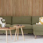 Un salon moderne dispose d'un élégant canapé vert, de deux tables en bois, de coussins décoratifs et d'une plante en pot sur un élégant panneau de bois, rappelant l'ambiance sophistiquée d'une salle d'exposition. Canape Paris by Atelier Bis