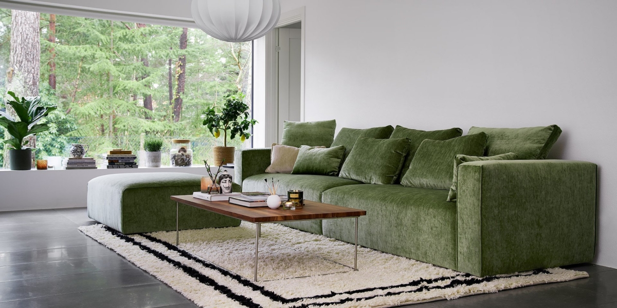 Salon moderne avec un canapé en velours vert chic, une table basse en bois et une grande fenêtre offrant une vue sur la verdure luxuriante à l'extérieur, rappelant un showroom parisien élégant. Canape Paris by Atelier Bis