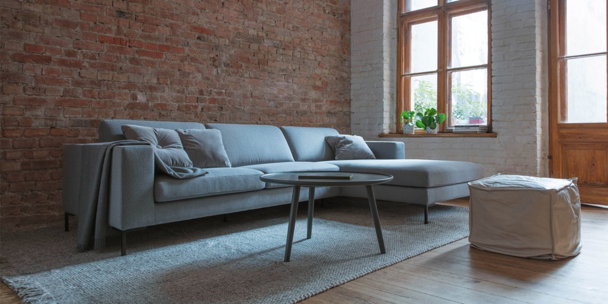 Un canapé gris et une table basse ronde ornent la pièce rustique avec un mur de briques, un parquet en bois et une grande fenêtre, offrant une touche de qualité parisienne. Canape Paris by Atelier Bis