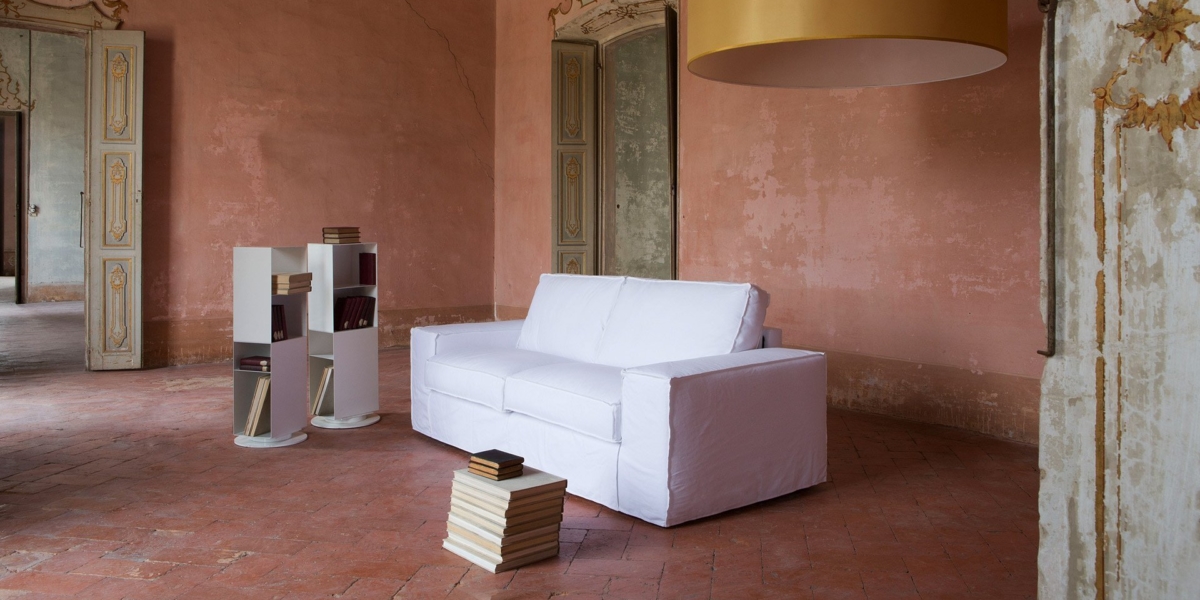 Chambre spacieuse de style parisien avec un canapé blanc, des étagères et une petite table remplie de livres, éclairée par une grande lampe suspendue, créant l'ambiance parfaite d'un showroom. Canape Paris by Atelier Bis