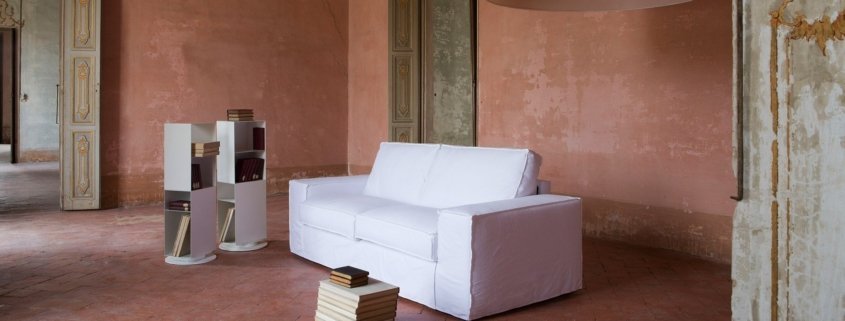 Chambre spacieuse de style parisien avec un canapé blanc, des étagères et une petite table remplie de livres, éclairée par une grande lampe suspendue, créant l'ambiance parfaite d'un showroom. Canape Paris by Atelier Bis