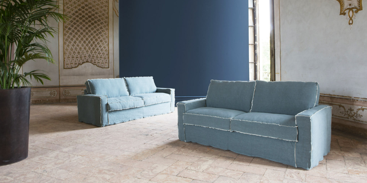 Deux canapés bleus dans une pièce élégante avec des murs à motifs, de grandes fenêtres et une plante en pot sur un sol en pierre dégagent un air de qualité parisienne. Canape Paris by Atelier Bis