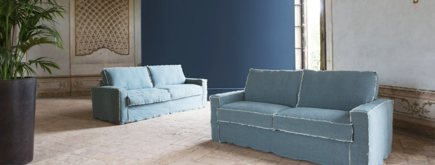Deux canapés bleus dans une pièce élégante avec des murs à motifs, de grandes fenêtres et une plante en pot sur un sol en pierre dégagent un air de qualité parisienne. Canape Paris by Atelier Bis