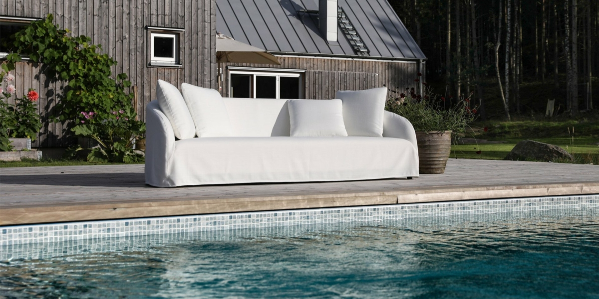Un canapé blanc orne la terrasse en bois au bord de la piscine, complété par une maison en bois moderne et un arrière-plan forestier serein, dégageant un sentiment de qualité qui ne serait pas déplacé à Paris. Canape Paris by Atelier Bis