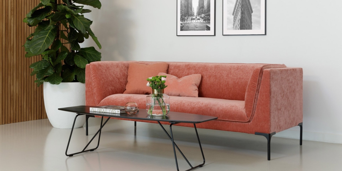 Dans la salle d'exposition, un canapé corail orné de coussins roses s'associe élégamment à une table basse noire et à une plante en pot, le tout adossé à un mur présentant des œuvres d'art encadrées. Canape Paris by Atelier Bis