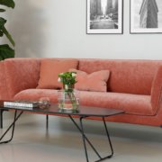 Dans la salle d'exposition, un canapé corail orné de coussins roses s'associe élégamment à une table basse noire et à une plante en pot, le tout adossé à un mur présentant des œuvres d'art encadrées. Canape Paris by Atelier Bis