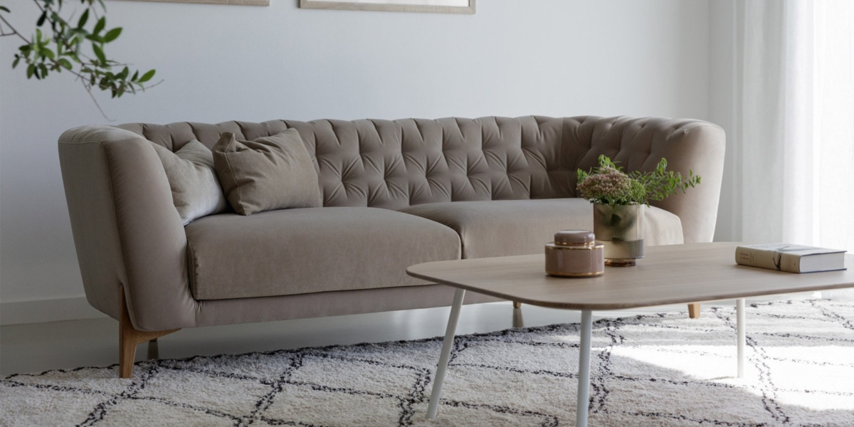 Un canapé capitonné beige chic avec des coussins est placé à côté d'une table basse en bois sur un tapis à motifs, créant un point focal élégant dans cette pièce lumineuse et minimaliste rappelant le charme parisien. Canape Paris by Atelier Bis