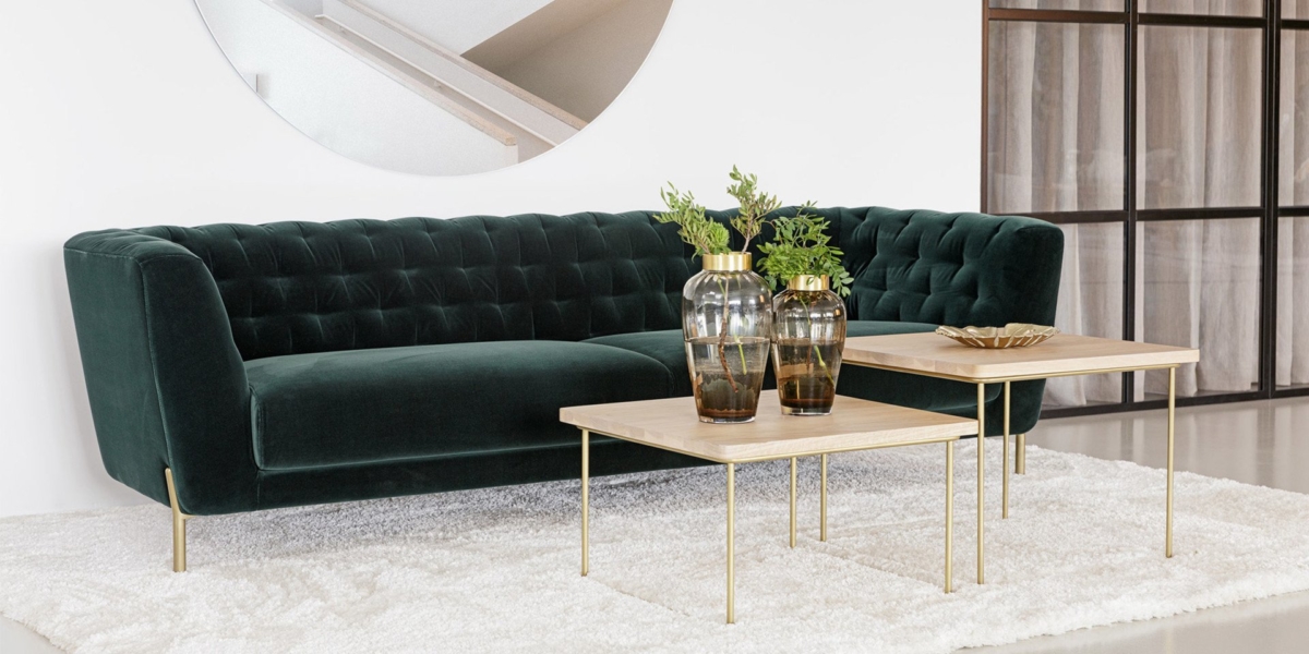 Un canapé en velours vert avec des plantes sur des tables en bois, posé élégamment sur un tapis blanc. Un miroir rond et une cloison en verre ornent le fond, offrant une touche de charme et de style parisien qui équilibre sans effort le prix et la qualité. Canape Paris by Atelier Bis