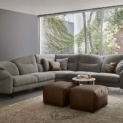 Salon confortable avec une décoration de qualité salle d'exposition, comprenant un grand canapé sectionnel gris, des poufs marron, de l'art abstrait et une vue invitante sur la verdure extérieure. Canape Paris by Atelier Bis