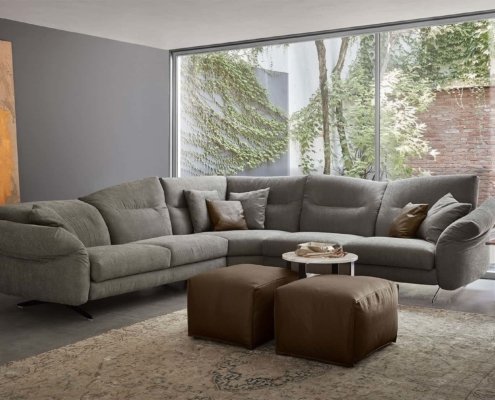 Salon confortable avec une décoration de qualité salle d'exposition, comprenant un grand canapé sectionnel gris, des poufs marron, de l'art abstrait et une vue invitante sur la verdure extérieure. Canape Paris by Atelier Bis