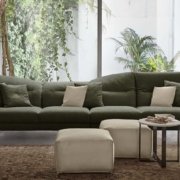 Une ambiance parisienne chic imprègne la pièce avec un canapé vert accompagné de coussins beiges, deux poufs blancs et une petite table ronde. De grandes fenêtres inondent l'espace de lumière, tandis que des plantes luxuriantes ajoutent une touche de verdure. Canape Paris by Atelier Bis