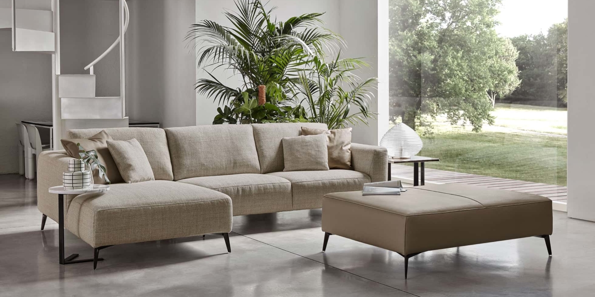 Un canapé beige moderne et un pouf ornent une salle d'exposition lumineuse, entourée de grandes fenêtres, de plantes vertes luxuriantes et d'un escalier en colimaçon saisissant. Canape Paris by Atelier Bis