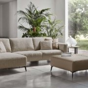 Un canapé beige moderne et un pouf ornent une salle d'exposition lumineuse, entourée de grandes fenêtres, de plantes vertes luxuriantes et d'un escalier en colimaçon saisissant. Canape Paris by Atelier Bis