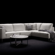 Canapé sectionnel blanc avec coussins et jeté gris, flanqué de tables rondes sur fond noir, reflétant l'élégance chic du décor parisien, où la qualité rencontre le style sans effort. Canape Paris by Atelier Bis