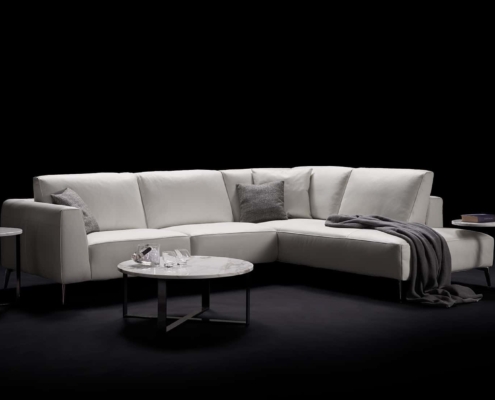 Canapé sectionnel blanc avec coussins et jeté gris, flanqué de tables rondes sur fond noir, reflétant l'élégance chic du décor parisien, où la qualité rencontre le style sans effort. Canape Paris by Atelier Bis