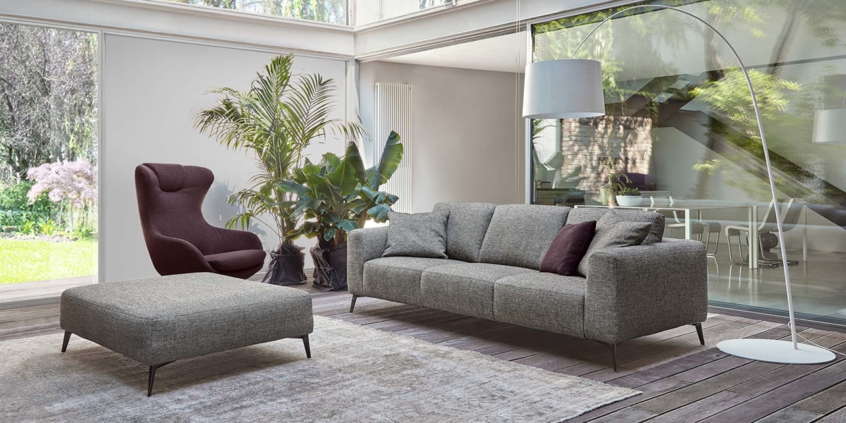 Découvrez la qualité d'un salon moderne avec un canapé gris épuré, complété par un pouf et un fauteuil. De grandes fenêtres inondent l'espace de lumière tandis que les plantes ajoutent une touche de nature, le tout éclairé par un élégant lampadaire. Canape Paris by Atelier Bis
