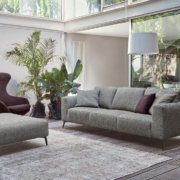 Découvrez la qualité d'un salon moderne avec un canapé gris épuré, complété par un pouf et un fauteuil. De grandes fenêtres inondent l'espace de lumière tandis que les plantes ajoutent une touche de nature, le tout éclairé par un élégant lampadaire. Canape Paris by Atelier Bis