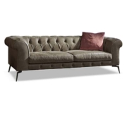 Ce canapé capitonné vert élégant, avec ses accoudoirs roulés et ses pieds élancés, respire le charme parisien. Un coussin marron unique ajoute une touche de sophistication sur le côté droit, mettant en valeur sa qualité exceptionnelle. Canape Paris by Atelier Bis