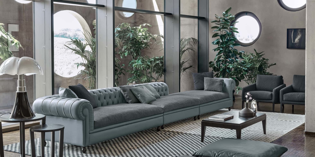 Un salon moderne à Paris dispose d'un long canapé gris, de fenêtres rondes, de plantes vertes et d'un tapis à carreaux, tous incarnant une qualité exquise. Canape Paris by Atelier Bis