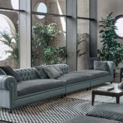 Un salon moderne à Paris dispose d'un long canapé gris, de fenêtres rondes, de plantes vertes et d'un tapis à carreaux, tous incarnant une qualité exquise. Canape Paris by Atelier Bis