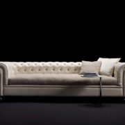 Élégant canapé capitonné de couleur crème avec des coussins beiges et une petite table contenant des livres, présenté dans notre salle d'exposition sur un fond noir, offrant une qualité inégalée à un prix imbattable. Canape Paris by Atelier Bis