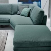 Dans une salle d'exposition élégante, un canapé sectionnel vert avec des coussins bleu sarcelle repose gracieusement sur un sol gris clair, accompagné d'un tapis chic et d'œuvres d'art partiellement visibles. Canape Paris by Atelier Bis