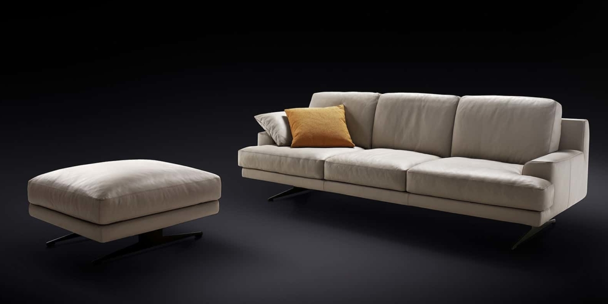 Canapé beige avec un coussin jaune et un pouf assorti présenté sur un fond noir, illustrant l'élégance de qualité salle d'exposition. Canape Paris by Atelier Bis