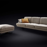 Canapé beige avec un coussin jaune et un pouf assorti présenté sur un fond noir, illustrant l'élégance de qualité salle d'exposition. Canape Paris by Atelier Bis