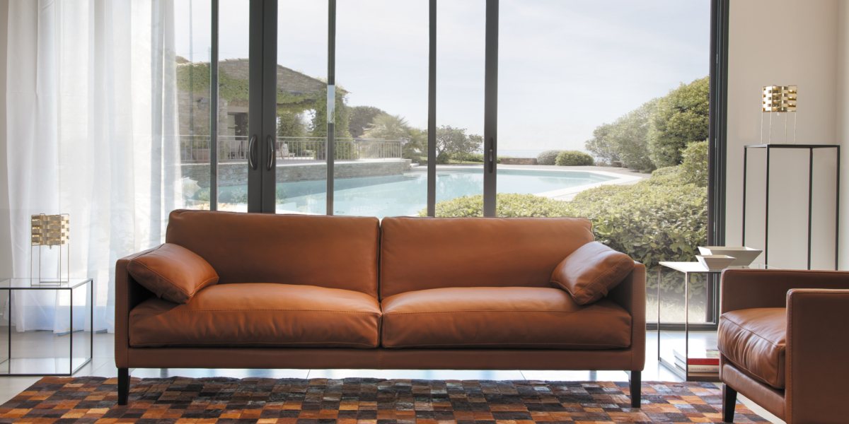 Un canapé en cuir marron dans un salon moderne présente une qualité exquise, avec une vue sur la piscine et la verdure luxuriante à travers des portes coulissantes en verre, offrant un cadre digne d'une salle d'exposition. Canape Paris by Atelier Bis
