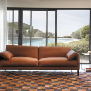 Un canapé en cuir marron dans un salon moderne présente une qualité exquise, avec une vue sur la piscine et la verdure luxuriante à travers des portes coulissantes en verre, offrant un cadre digne d'une salle d'exposition. Canape Paris by Atelier Bis