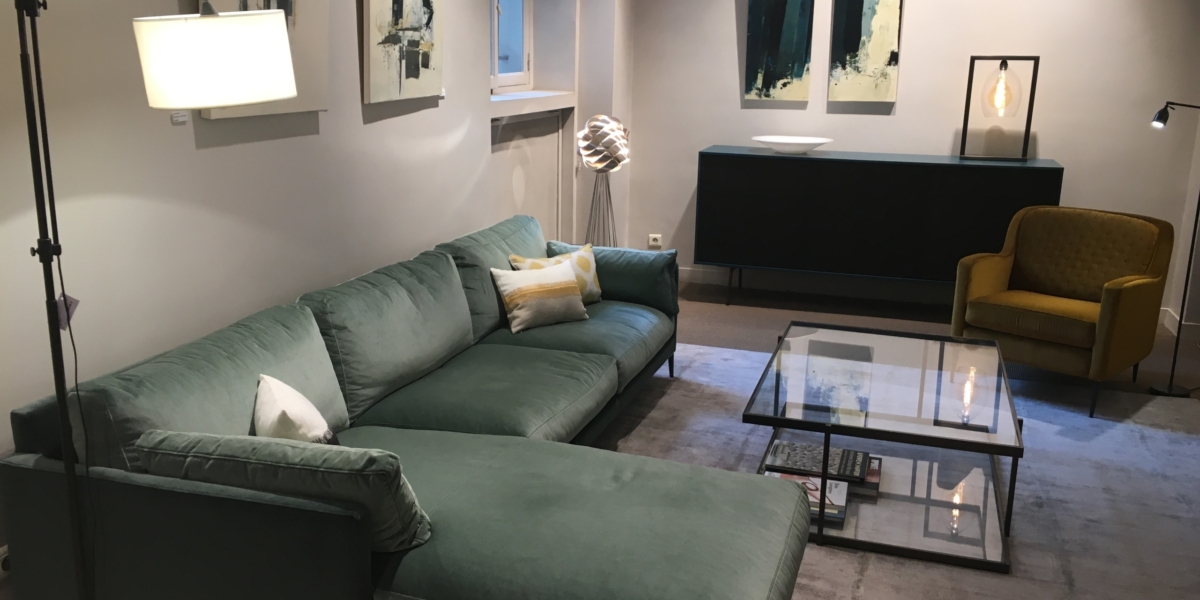 Salon moderne aux allures parisiennes : un canapé sectionnel vert, des œuvres d'art abstraites et une élégante table basse en verre posée sur un tapis gris, rappelant une élégante vitrine d'exposition. Canape Paris by Atelier Bis