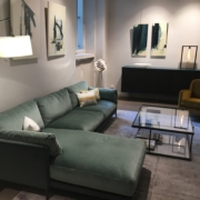 Salon moderne aux allures parisiennes : un canapé sectionnel vert, des œuvres d'art abstraites et une élégante table basse en verre posée sur un tapis gris, rappelant une élégante vitrine d'exposition. Canape Paris by Atelier Bis