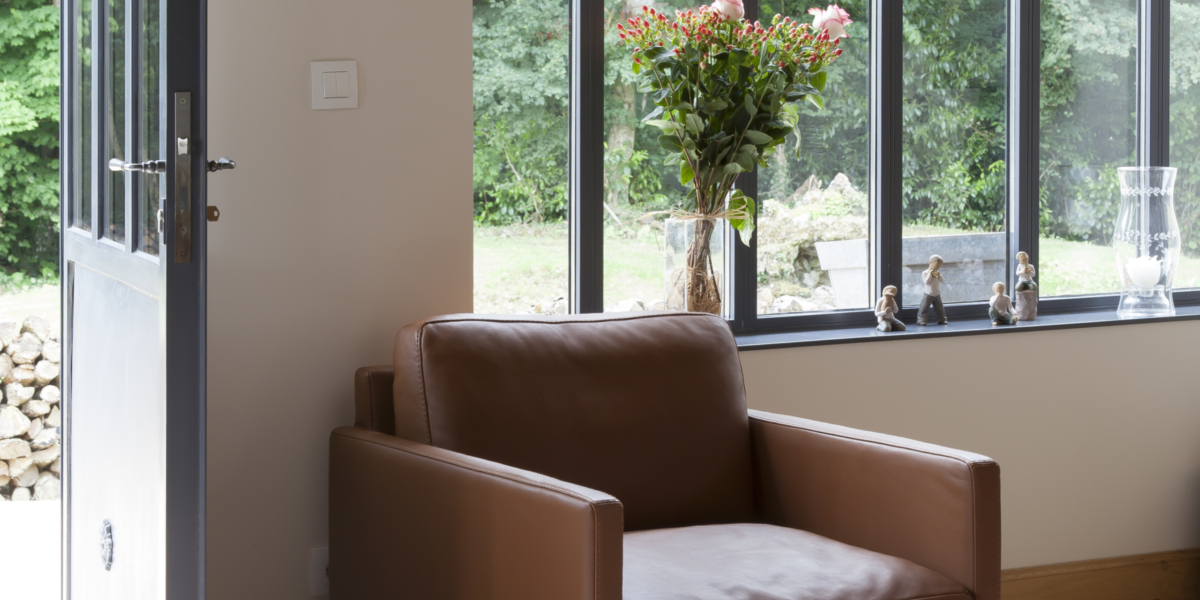 Un fauteuil marron trône près d'une porte vitrée, accompagné de fleurs sur une table près de grandes fenêtres donnant sur une verdure luxuriante, offrant une scène d'une qualité inégalée. Canape Paris by Atelier Bis