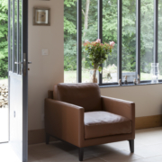 Un fauteuil marron trône près d'une porte vitrée, accompagné de fleurs sur une table près de grandes fenêtres donnant sur une verdure luxuriante, offrant une scène d'une qualité inégalée. Canape Paris by Atelier Bis