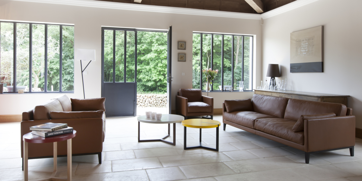 Salon moderne avec canapés en cuir, tables rondes, grandes fenêtres et vue sur la verdure extérieure, présentant un design de qualité showroom à un prix imbattable. Canape Paris by Atelier Bis