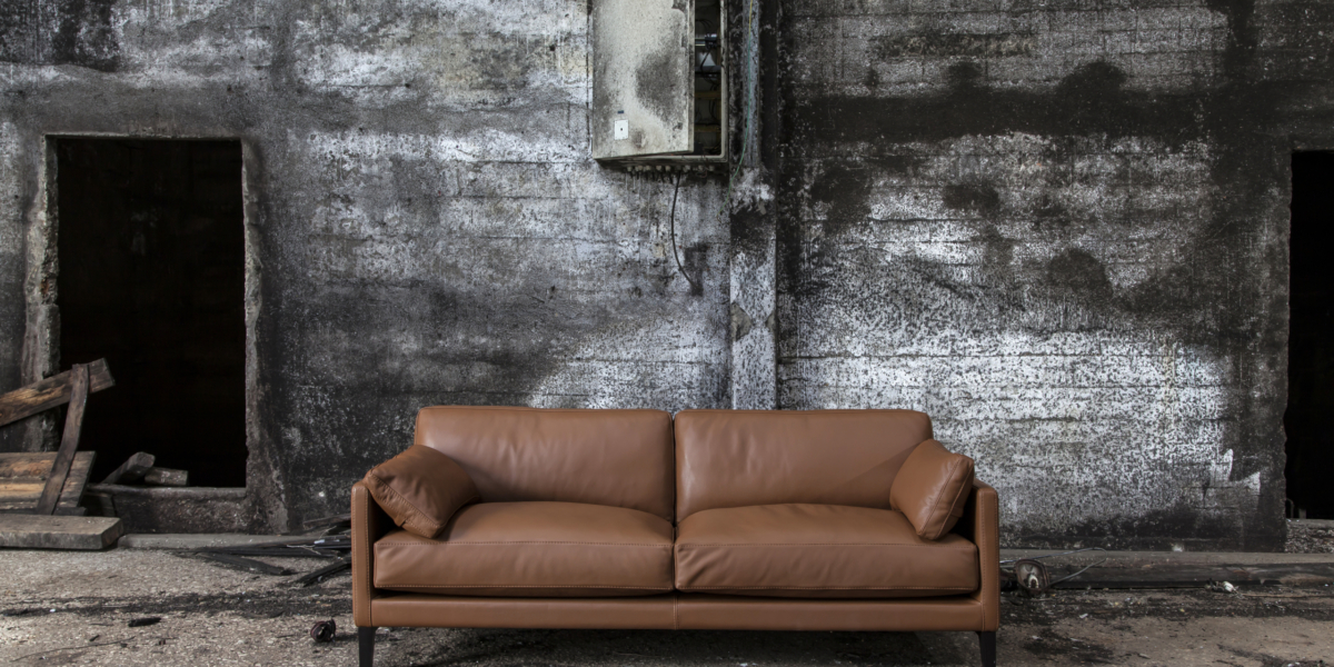 Un canapé en cuir marron trône majestueusement devant un mur sombre et patiné avec deux portes ouvertes, incarnant le mélange parfait de prix et de qualité, directement depuis le showroom. Canape Paris by Atelier Bis