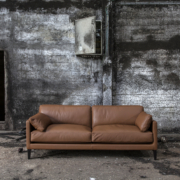 Un canapé en cuir marron trône majestueusement devant un mur sombre et patiné avec deux portes ouvertes, incarnant le mélange parfait de prix et de qualité, directement depuis le showroom. Canape Paris by Atelier Bis