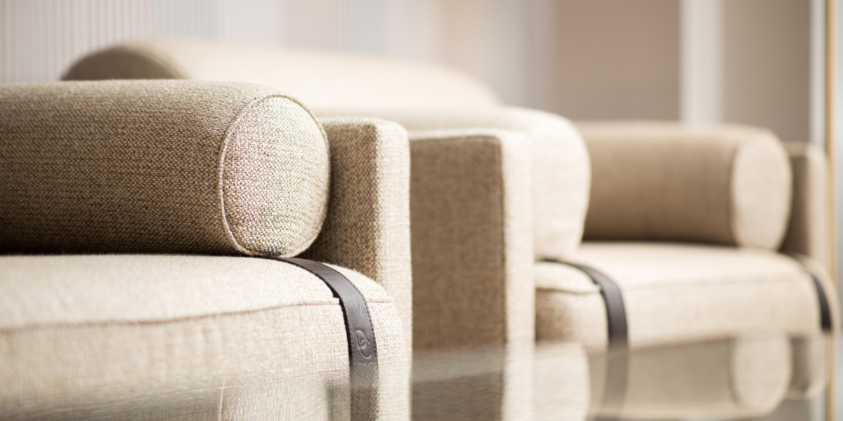 Gros plan de deux canapés beiges aux accoudoirs cylindriques et aux accents métalliques, rappelant une vitrine d'exposition haut de gamme, se reflétant sur une surface en verre. Canape Paris by Atelier Bis