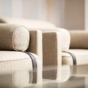 Gros plan de deux canapés beiges aux accoudoirs cylindriques et aux accents métalliques, rappelant une vitrine d'exposition haut de gamme, se reflétant sur une surface en verre. Canape Paris by Atelier Bis