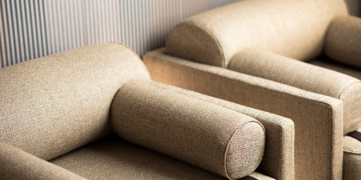 Deux fauteuils beiges rembourrés avec accoudoirs cylindriques se dressent élégamment sur un fond mural texturé, rappelant une vitrine d'exposition chic. Canape Paris by Atelier Bis