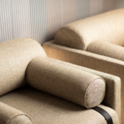 Deux fauteuils beiges rembourrés avec accoudoirs cylindriques se dressent élégamment sur un fond mural texturé, rappelant une vitrine d'exposition chic. Canape Paris by Atelier Bis