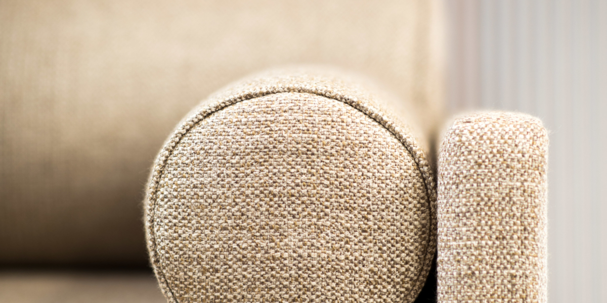 Gros plan d'un accoudoir de canapé rembourré beige, mettant en valeur la qualité de son coussin cylindrique et de sa sangle d'accent sombre contre un mur rayé, rappelant une salle d'exposition élégante. Canape Paris by Atelier Bis