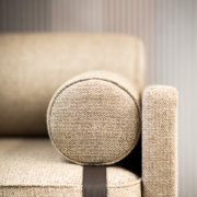 Gros plan d'un accoudoir de canapé rembourré beige, mettant en valeur la qualité de son coussin cylindrique et de sa sangle d'accent sombre contre un mur rayé, rappelant une salle d'exposition élégante. Canape Paris by Atelier Bis