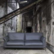 Un canapé gris foncé dans un décor industriel patiné avec des poutres métalliques et des murs en béton texturé, rappelant un showroom parisien chic. Canape Paris by Atelier Bis