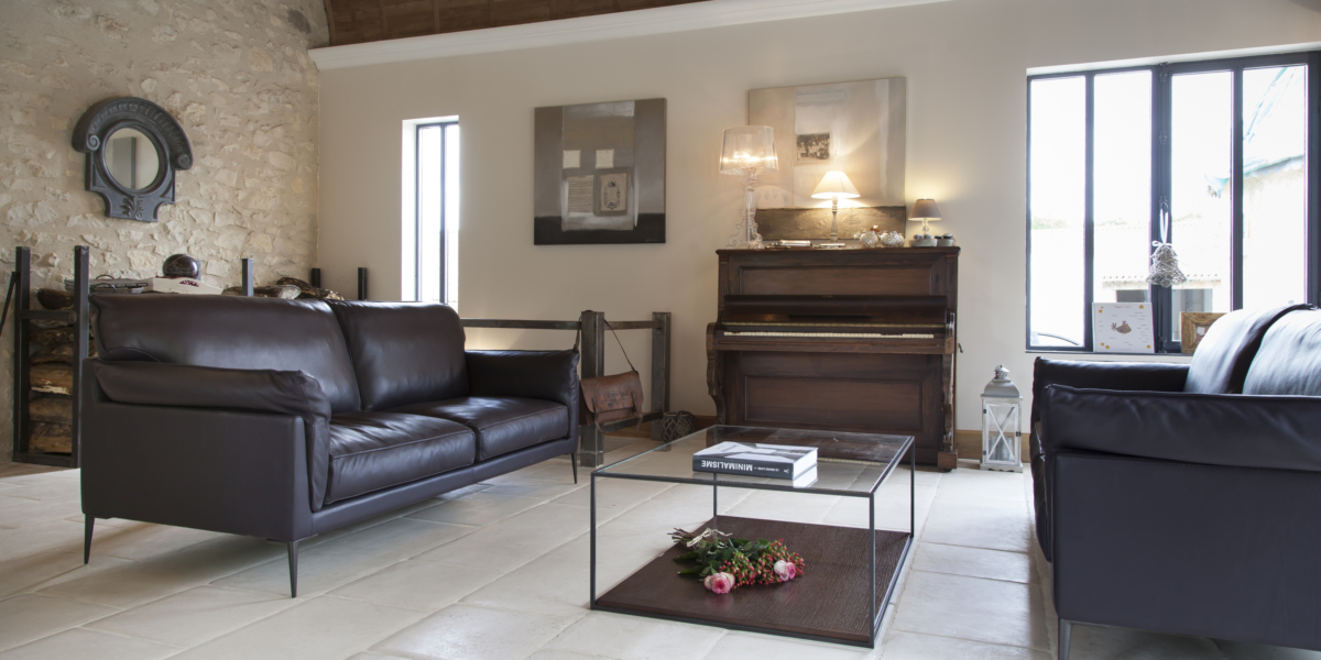 Un salon moderne est doté de deux canapés noirs rappelant l'élégance d'une salle d'exposition, complétés par une élégante table basse en verre et un piano. Le mur d'accent en pierre ajoute une touche de qualité, rendant cet espace à la fois élégant et accueillant. Canape Paris by Atelier Bis