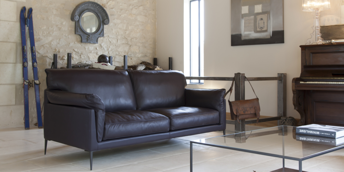 Un salon chic comprend un canapé en cuir foncé, une table en verre, un piano, des skis vintage et un mur en pierre rustique, rappelant un showroom parisien chaleureux. Canape Paris by Atelier Bis