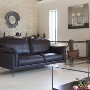 Un salon chic comprend un canapé en cuir foncé, une table en verre, un piano, des skis vintage et un mur en pierre rustique, rappelant un showroom parisien chaleureux. Canape Paris by Atelier Bis