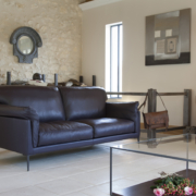 Salon moderne avec canapé en cuir foncé, table basse en verre et piano, rappelant un showroom parisien. L'art mural complète les skis décoratifs contre un mur en pierre. Canape Paris by Atelier Bis