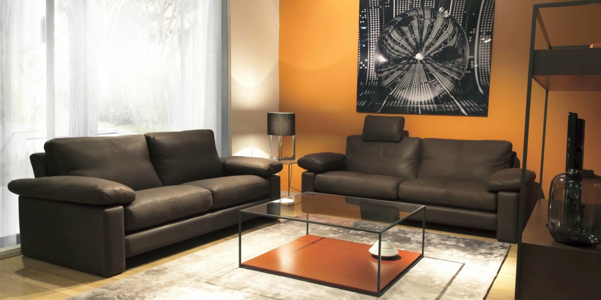 Un salon chic et moderne comprend deux canapés marron, une table basse en verre et des œuvres d'art murales abstraites sur un mur orange, rappelant un showroom parisien. Un lampadaire élégant complète l'ambiance élégante. Canape Paris by Atelier Bis