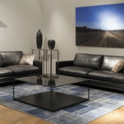 Découvrez un salon moderne où la qualité rencontre le style, avec des canapés noirs épurés, une table basse en verre et un tapis bleu en dessous. Un grand tableau de paysage orne le mur, créant une ambiance digne de tout showroom haut de gamme. Canape Paris by Atelier Bis